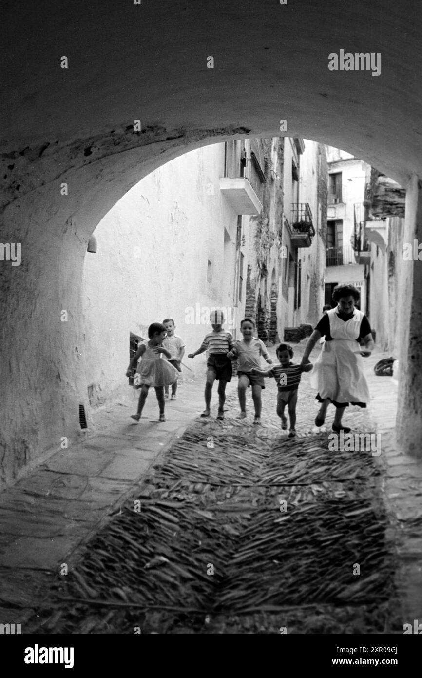 Fünf kleine Kinder laufen lachend eine Gasse in der Altstadt von Cadaqués hinab, dabei ist eine Frau, die ihrem weißen Kittel nach urteilen das Kindermädchen sein könnte und das jüngste Kind am Arm festhält, Costa Brava 1957. Fünf kleine Kinder laufen lachend durch eine Gasse in der Altstadt von Cadaqués, mit einer Frau, die nach ihrem weißen Mantel die Kindermädchen sein könnte, die das jüngste Kind am Arm hält, Costa Brava 1957. Stockfoto