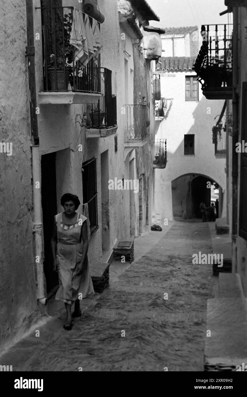 Eine Frau mit einer fleckigen Schürze über ihrem Kleid läuft mit gesenktem Blick eine Gasse in der Altstadt von Cadaqués hinauf, über ihr ragen ortstypische kleine Balkone aus den Häusern, Costa Brava 1957. Eine Frau mit einer befleckten Schürze über ihrem Kleid geht mit den Augen nach unten in einer Gasse in der Altstadt von Cadaqués, kleine Balkone, die typisch für die Gegend sind, ragen aus den Häusern über ihr hervor, Costa Brava 1957. Stockfoto