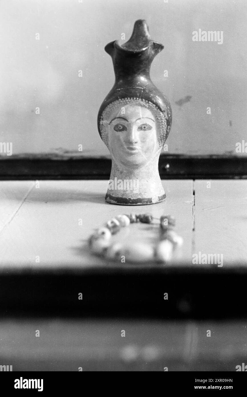 Kopfgefäße wie dieses in Form eines Frauenkopfes sind bevorzugt in der attischen Keramik zu finden, dieses weist schwarzfigurige Malerei und einen Kleeblatt-förmigen Ausguss auf und stammt vermutlich aus dem fünften Jahrhundert vor Christus, im Vordergrund des Bildes ist ein Perlenarmband zu erkennen, Empúries 1957. Kopfgefäße wie diese in Form eines Frauenkopfes finden sich meist in attischen Töpferwaren, diese mit schwarzfiguriger Malerei und einem kleeblattförmigen Auslauf und stammt wahrscheinlich aus dem 5. Jahrhundert v. Chr., ein perlenbesetztes Armband ist im Vordergrund des Bildes zu sehen, Empúries 1957. Stockfoto