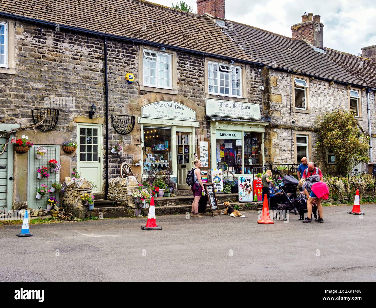 Castleton Derbyshire Vereinigtes Königreich. Malerische Dorfstraße mit Steinhäusern, Blumendekor und Menschen, die unter bewölktem Himmel mit Hunden interagieren. Stockfoto