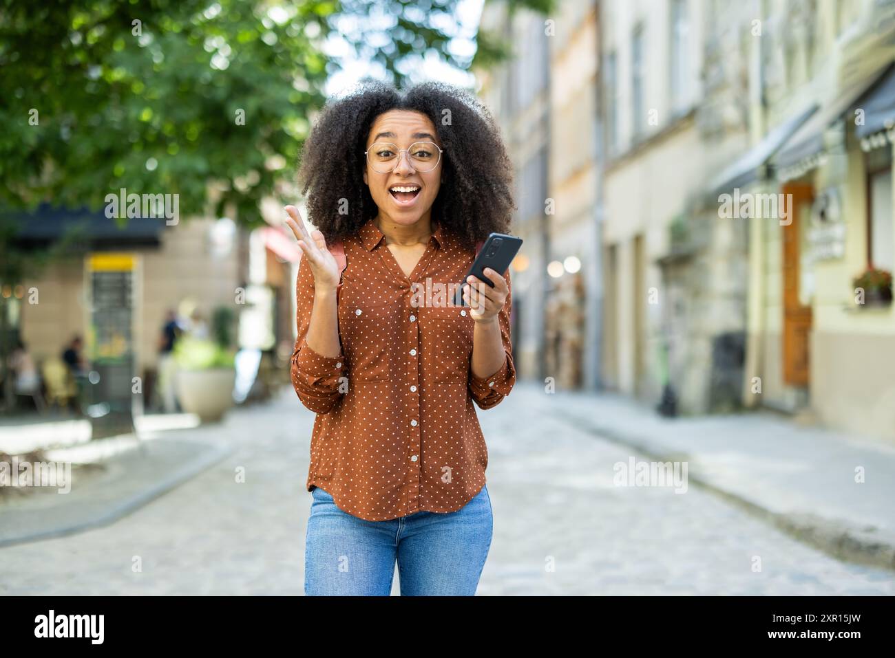 Fröhliche Frau mit lockigem Haar, Brille und lässigem Outfit, hält ein Telefon und feiert einen Sieg im Freien. Sie steht auf einer kopfsteingepflasterten Straße mit Gebäuden und Grün im Hintergrund. Stockfoto