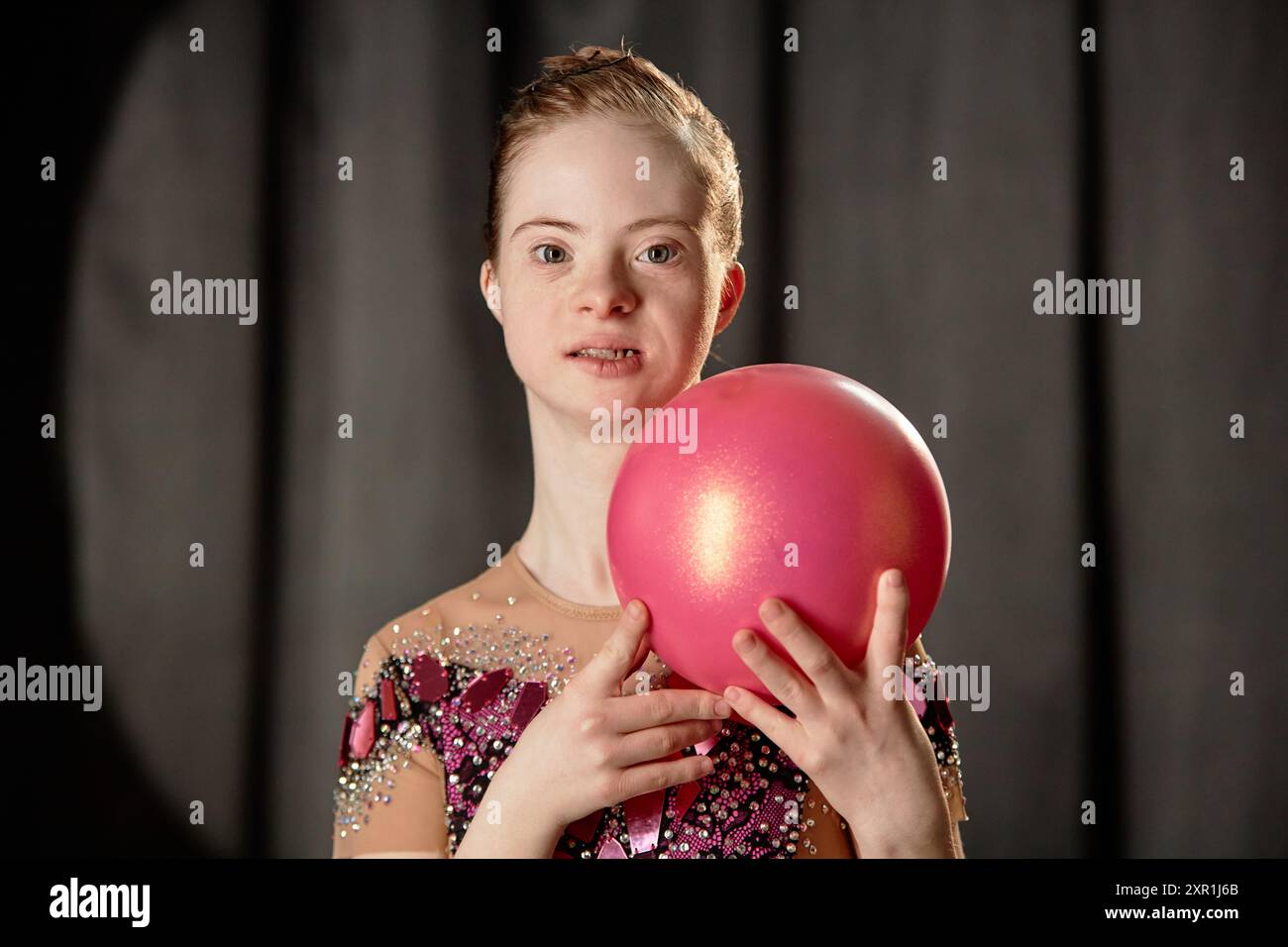 Porträt einer jungen Frau mit heller Haut und rosa Outfit, die einen glänzenden rosafarbenen Ball hält. Sie hat ein ausdrucksstarkes Gesicht und steht vor einem dunklen, verschwommenen Hintergrund, was einen auffälligen Kontrast schafft Stockfoto