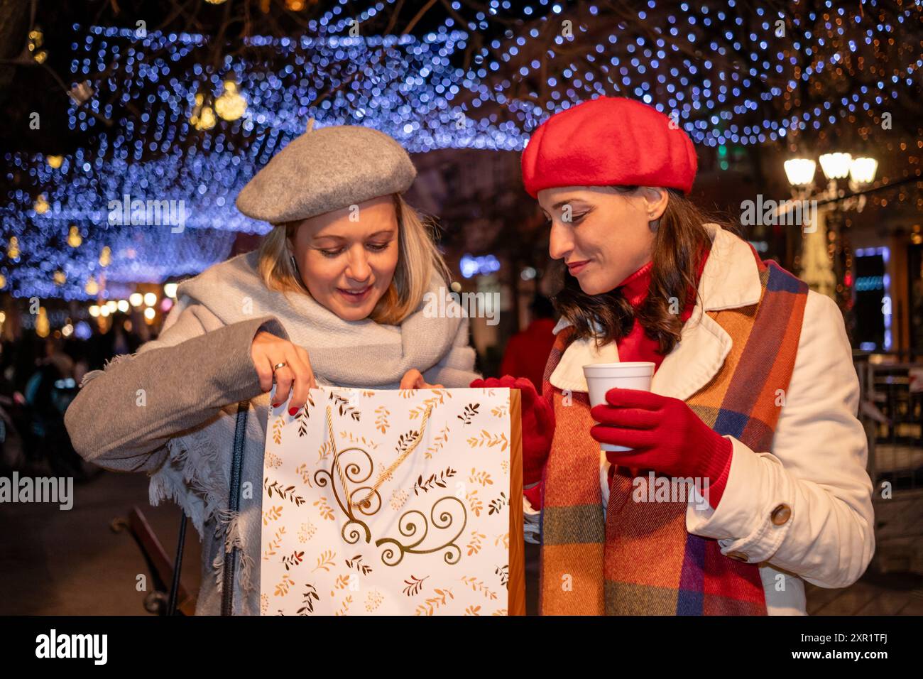 Freundinnen in Mänteln und französische Baskenmützen mit Einkaufstaschen genießen fröhliche Weihnachtseinkäufe und ein heißes Getränk auf einem festlich geschmückten Marktplatz. Stockfoto