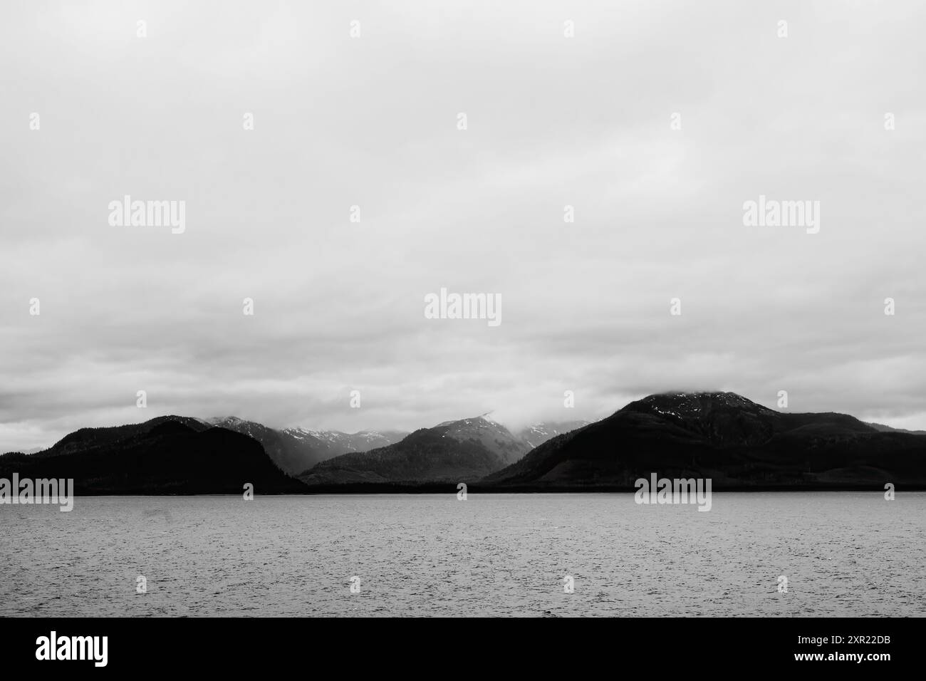 Grauer Himmel über dem rauen Gelände Alaskas und dem Wasser. Dunkle schattige Berge mit Schnee. Stockfoto