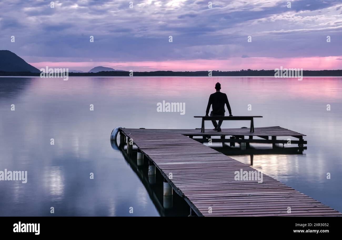 Mann, der auf einer hölzernen Pier-Bank sitzt und den Sonnenaufgang in Lagoa da Conceicao in Florianopolis - Brasilien beobachtet Stockfoto