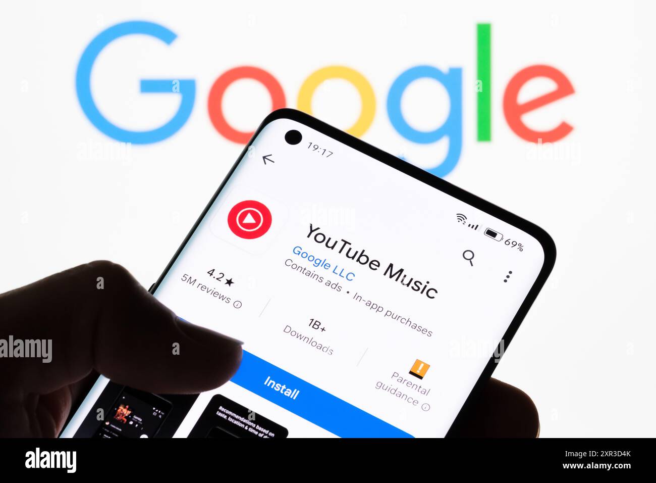 Nahaufnahme eines Smartphone-Bildschirms, auf dem die YouTube Music App mit dem Google-Logo im Hintergrund angezeigt wird, um die Verbindung zwischen YouTube Music und zu veranschaulichen Stockfoto