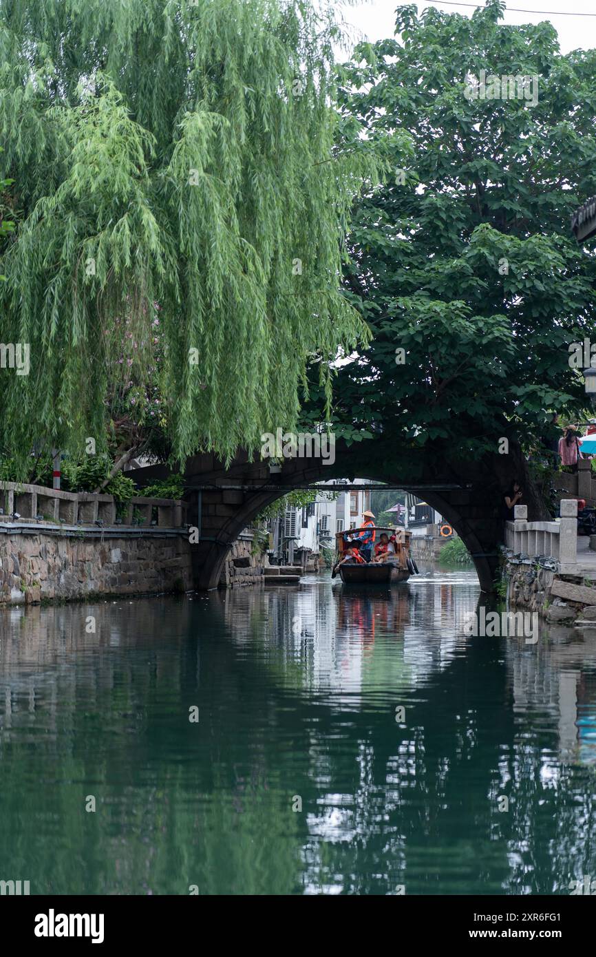 Suzhou, China - 11. Juni 2024 : Ein Boot mit Passagieren fährt unter einer Steinbogenbrücke, flankiert von üppigen grünen Bäumen und reflektiert in der Ruhe Stockfoto