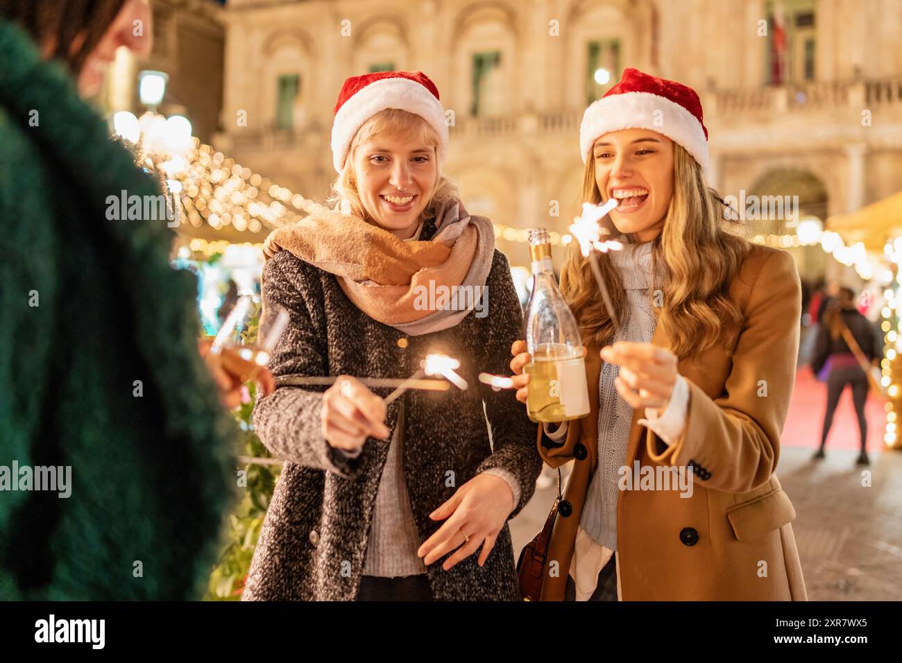 Fröhliche junge Frauen in Weihnachtsmützen feiern auf einem festlichen Weihnachtsmarkt. Das Bild fängt die Freude an der Weihnachtszeit mit glitzernden Lichtern, warmen Getränken, Stockfoto