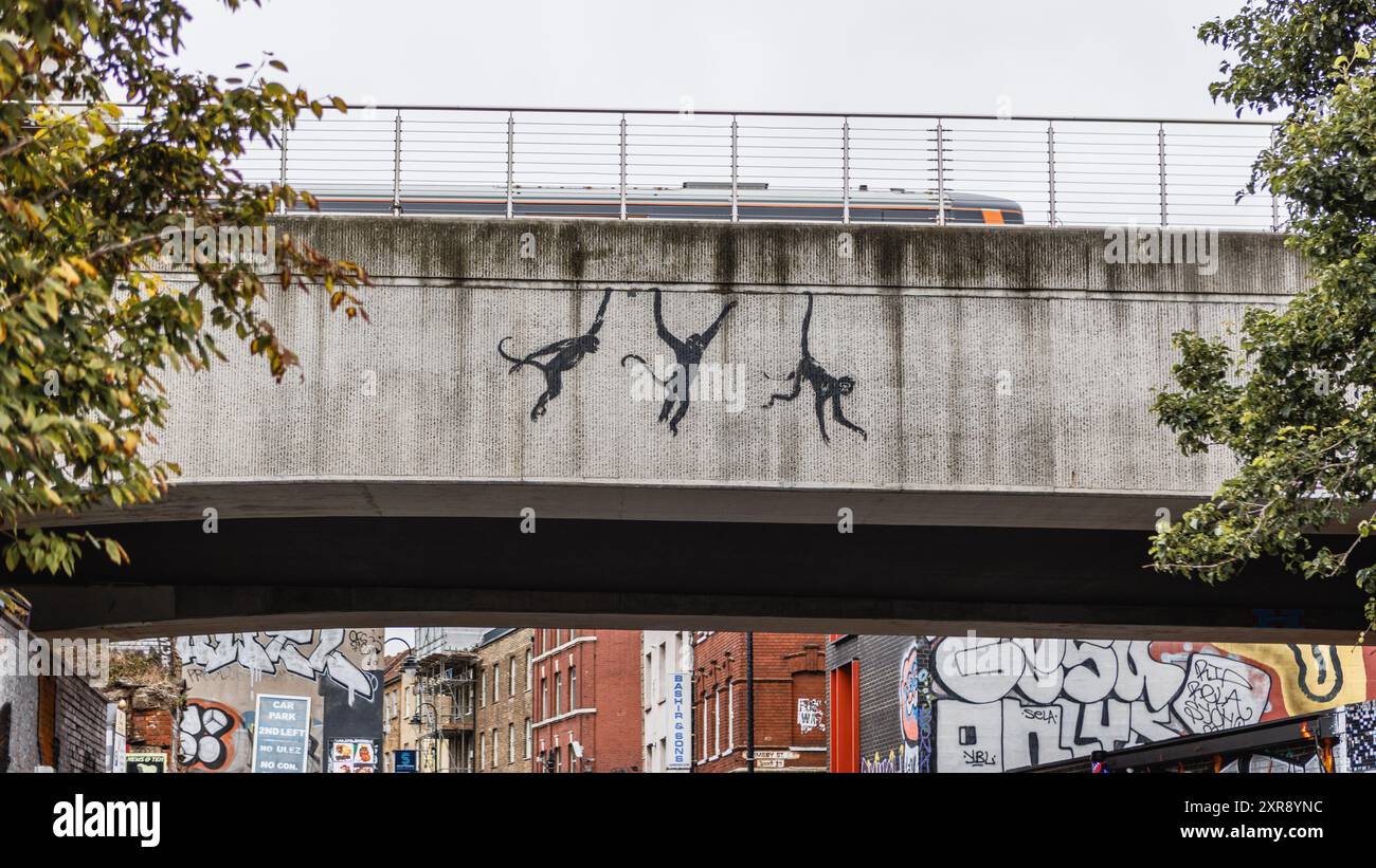 Banksys Londoner Tierserie: Drei schwingende Affen auf einer Eisenbahnbrücke auf der Brick Lane in Shoreditch, während ein Zug vorbeifährt. Stockfoto Banksys Londoner Tierserie: Drei schwingende Affen auf einer Eisenbahnbrücke auf der Brick Lane in Shoreditch, während ein Zug vorbeifährt. Stockfoto