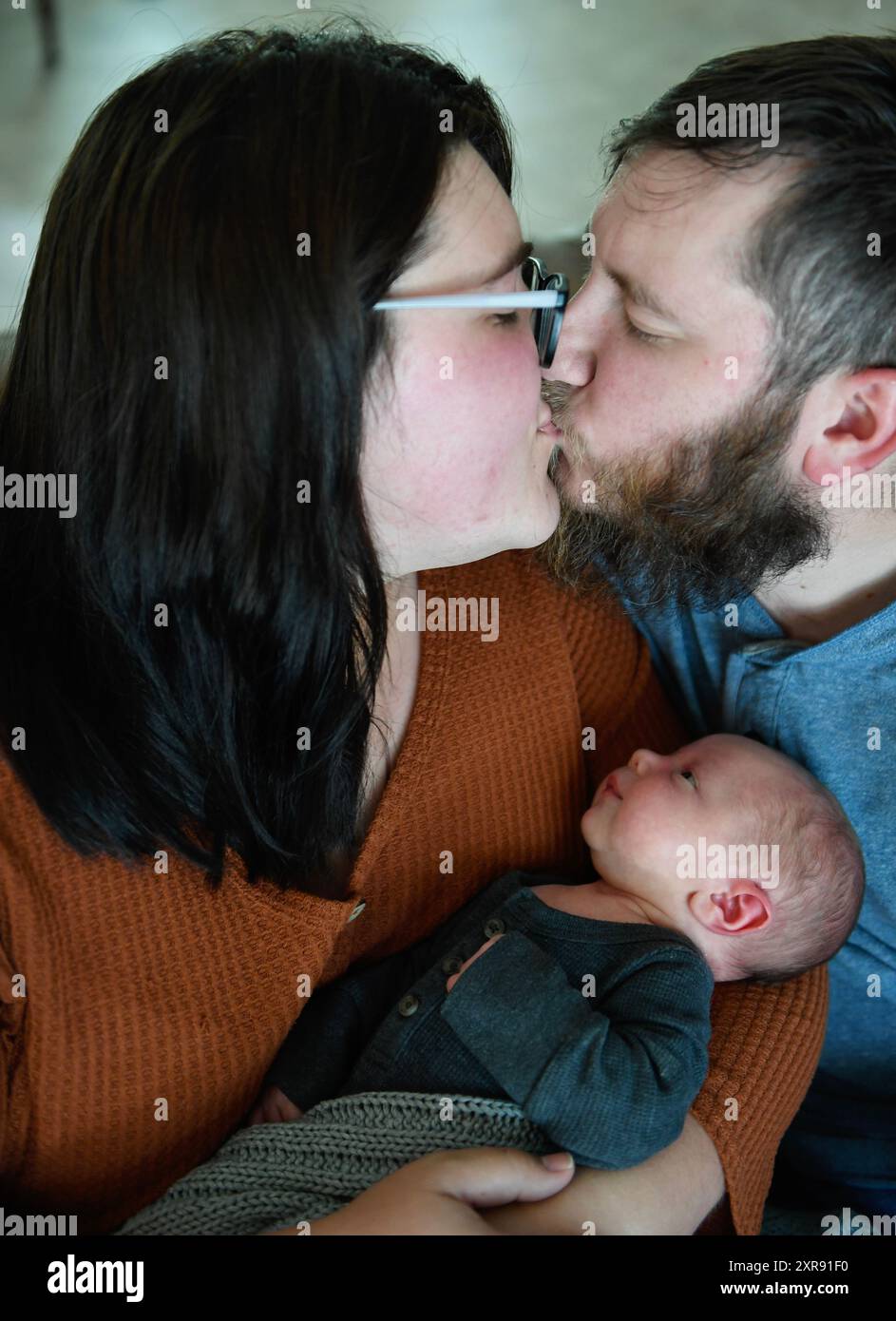 Eltern küssen sich, während sie ihr Neugeborenes in grauem Outfit halten. Stockfoto