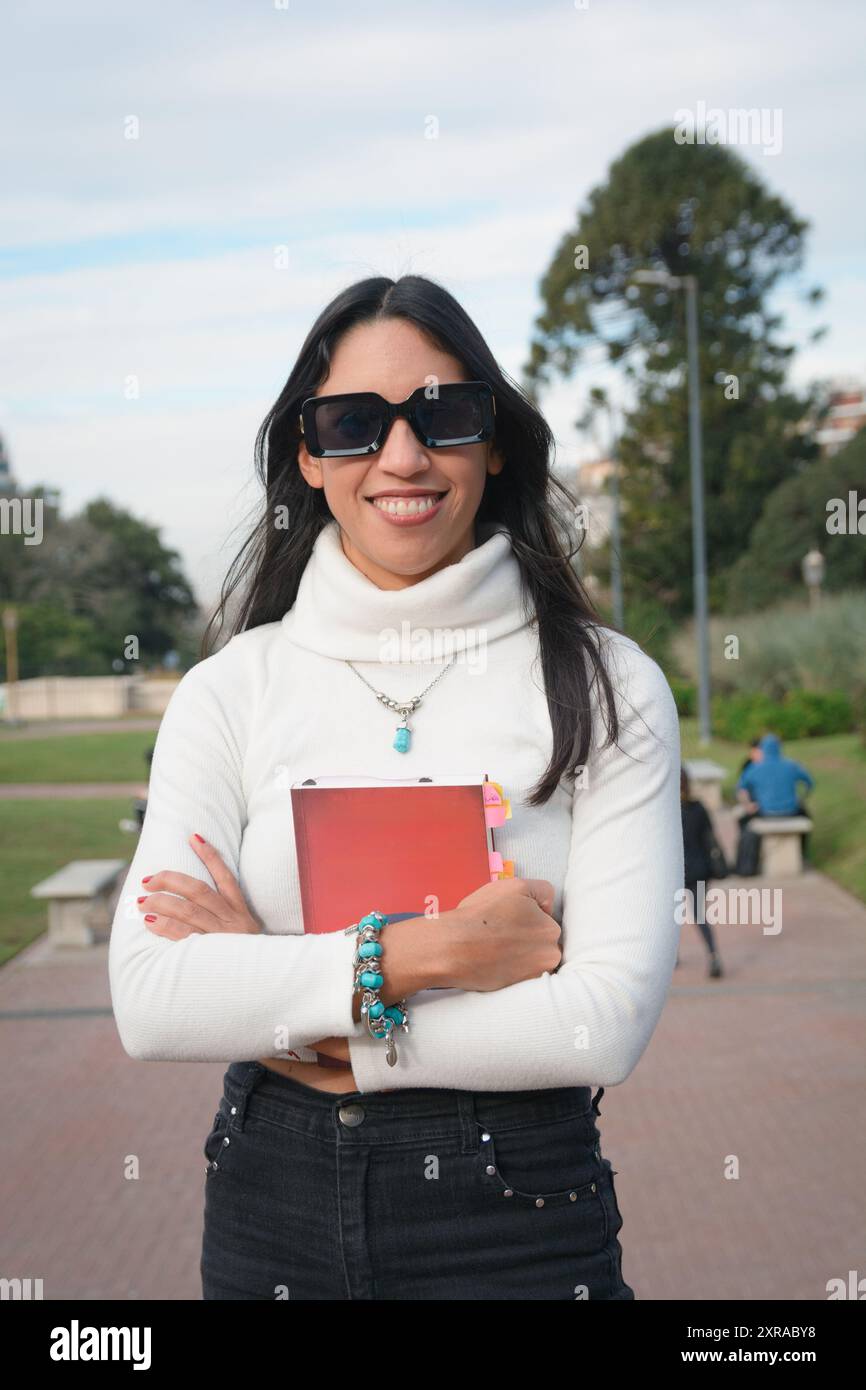 Porträt einer glücklichen jungen College-Frau aus argentinischer lateinischer Ethnie draußen, lächelnd und mit Sonnenbrille, mit guter Einstellung, umarmt ein Buch und Lo Stockfoto