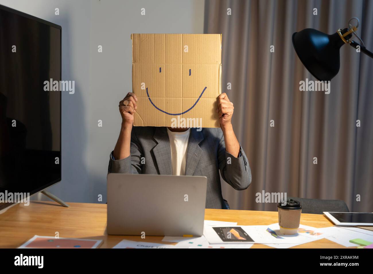 Ein junger Geschäftsmann hält einen Karton mit einem Smiley-Gesicht vor seinem Kopf, mit einer Pappschachtel auf dem Kopf, mit einem Smiley-Gesicht. Stockfoto