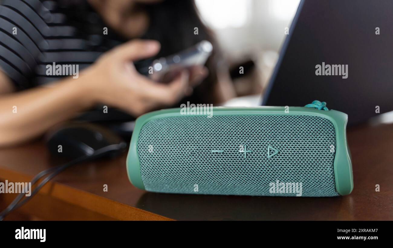 Nahaufnahme eines eleganten grünen Bluetooth-Lautsprechers auf einem Tisch zu Hause. Die Frau im Hintergrund sitzt an einem Tisch und arbeitet mit einem Laptop und einem sm Stockfoto