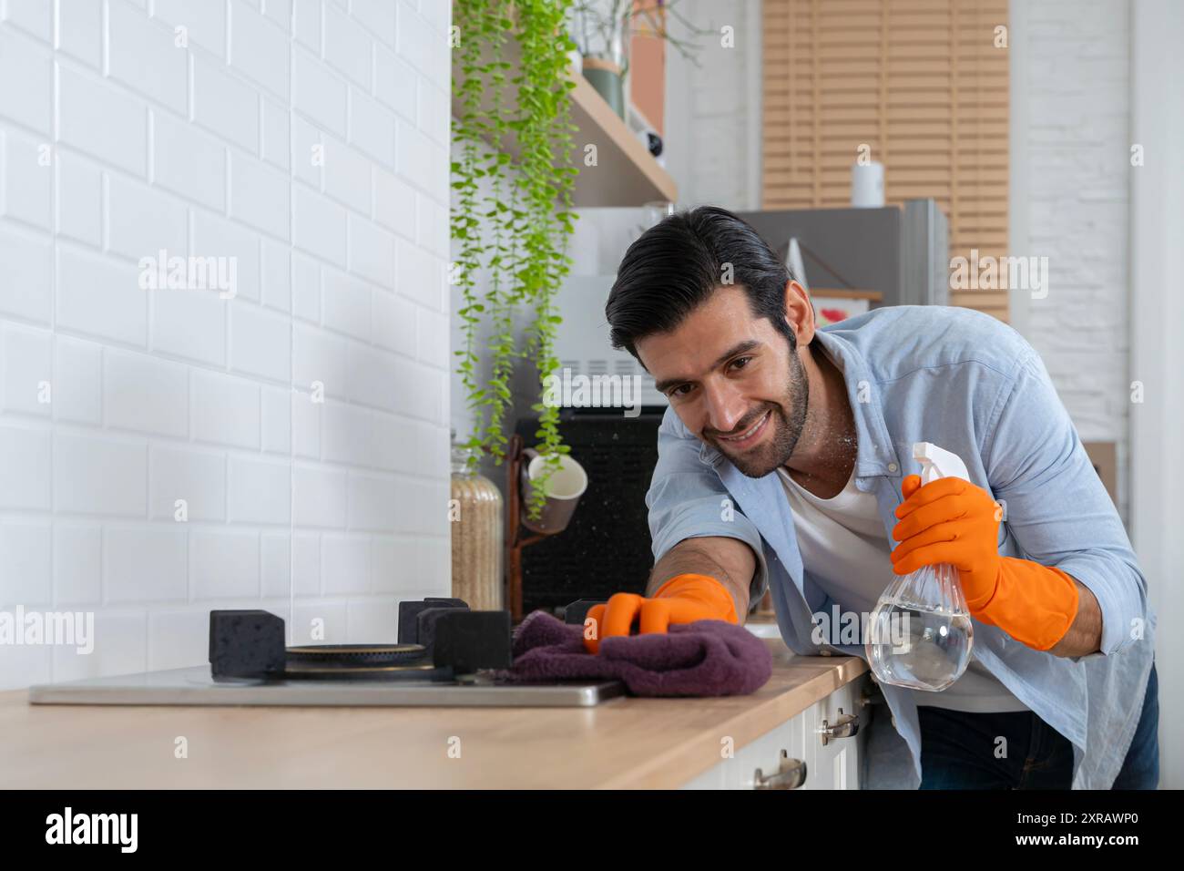 Junger gutaussehender Mann mit Schürze und Gummihandschuhen, der Holztisch in der Küche abwischt. Reinigungsservice-Konzept. Esstisch mit Tuch abwischen und lächeln. Stockfoto