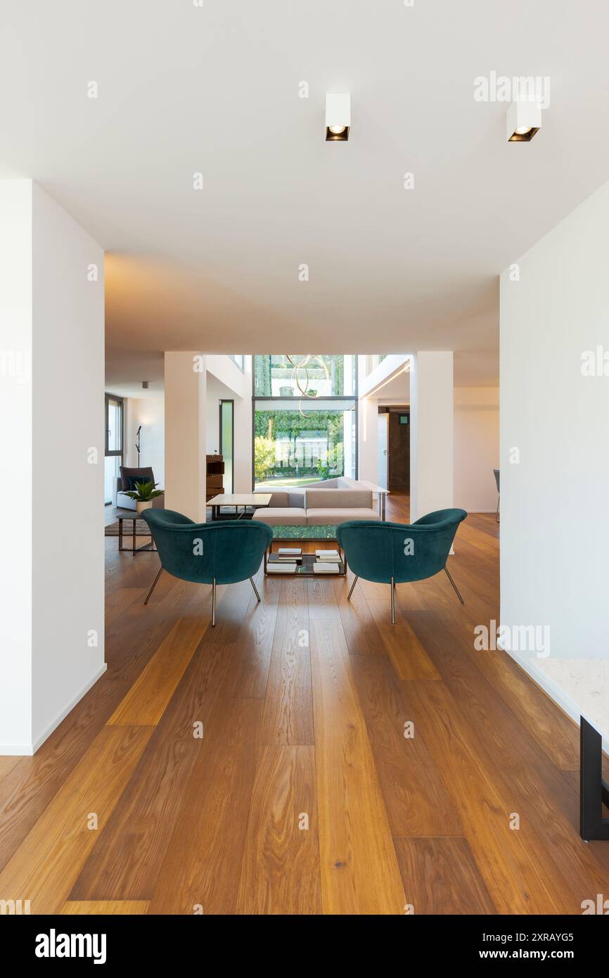 Modernes Wohnzimmer mit zwei türkisfarbenen Sesseln, einem Couchtisch und einem Sofa mit Kissen. Große Fenster mit Blick auf den Garten. Niemand drinnen Stockfoto