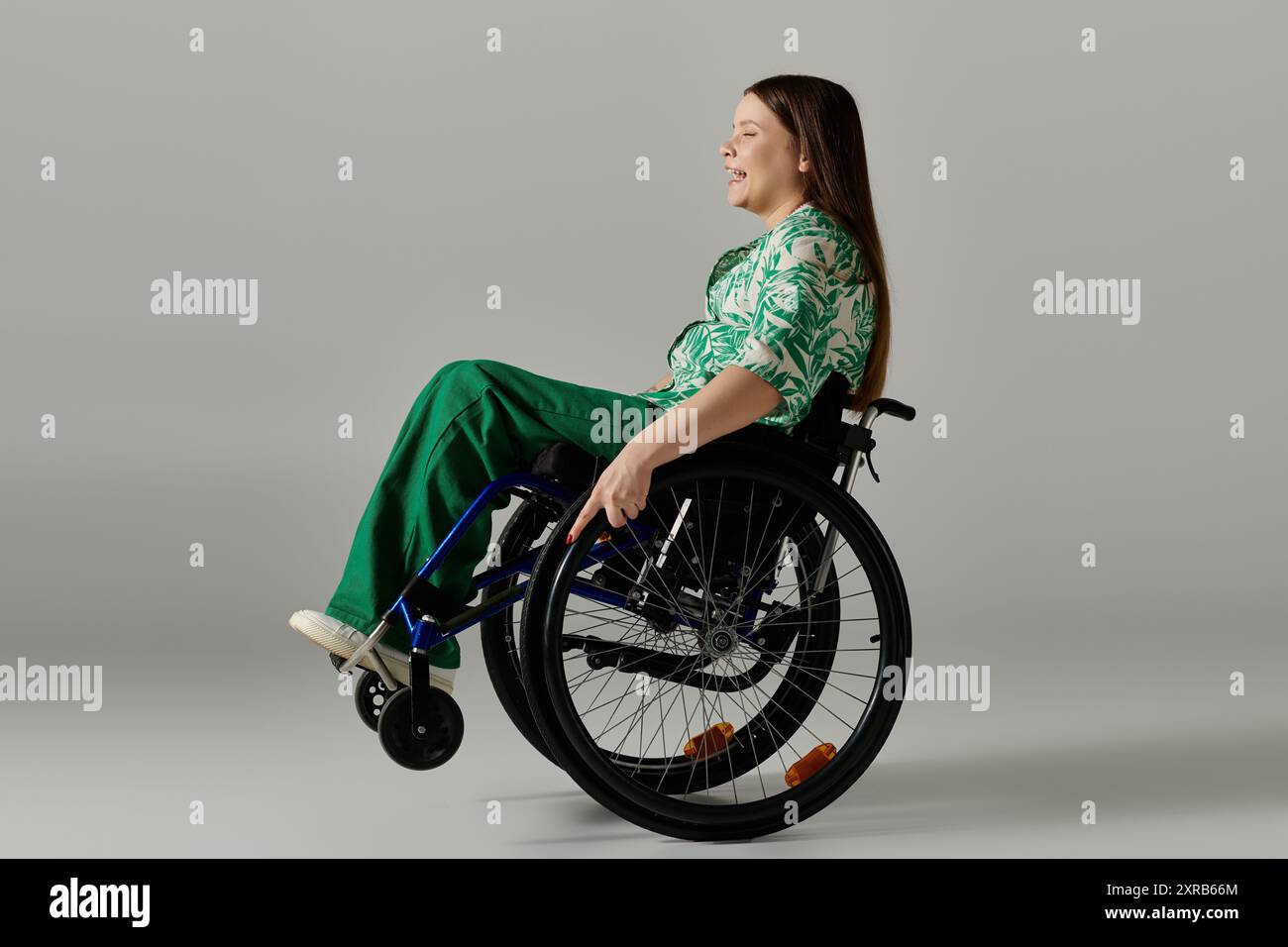 Eine junge Frau in grüner Kleidung sitzt in einem Rollstuhl vor grauem Hintergrund und lächelt hell. Stockfoto