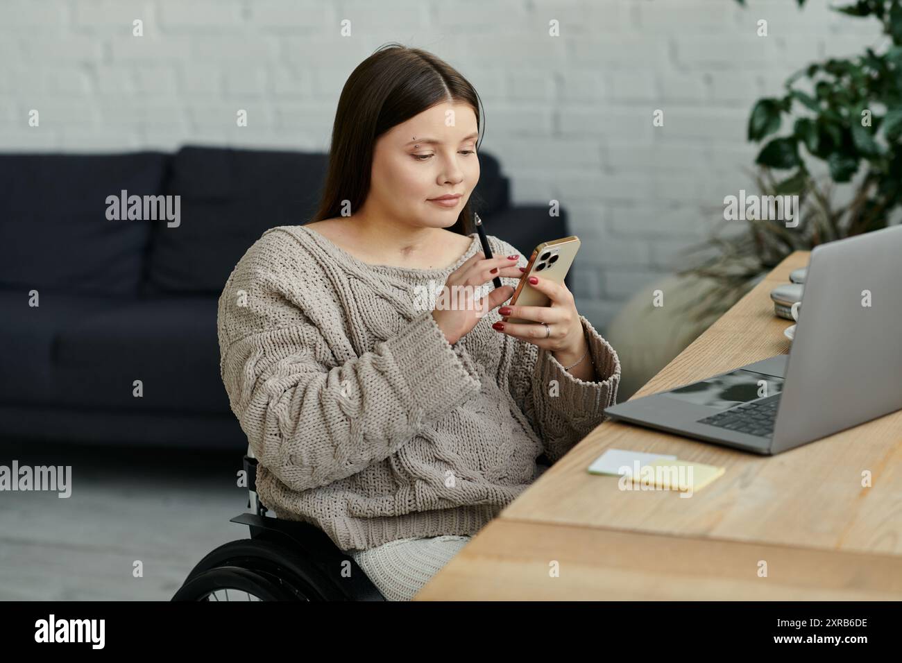 Eine junge Frau im Rollstuhl sitzt mit ihrem Smartphone an einem Tisch in ihrem Haus. Stockfoto