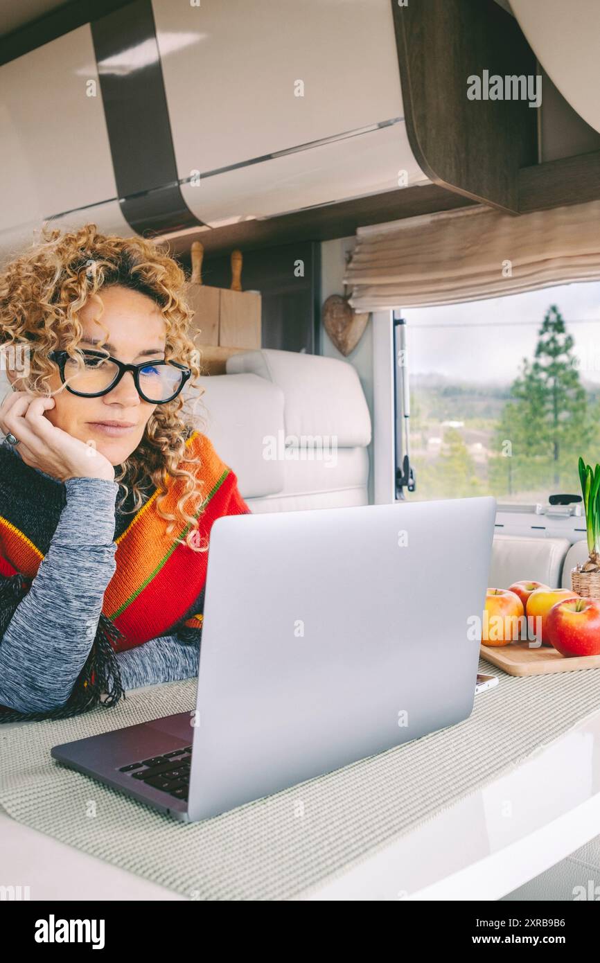 Das Leben von Transportern und der Lebensstil digitaler Nomaden. Niedliche, moderne Frau, die in einem Wohnmobil an einem Laptop arbeitet, mit Natur und Freiheit, malerischem Ort vor dem Fenster. Smart Working People Life Stockfoto