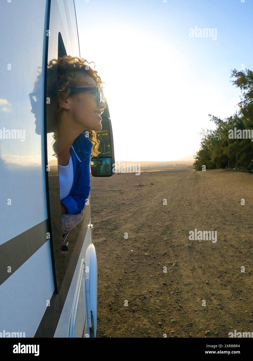 Blonde Frau mit lockigen Haaren schaut aus dem Fenster des sich bewegenden Campers. Das Weibchen schmeckt den Wind in ihrem Gesicht und lächelt. Reise- und Tourismuskonzept. Stockfoto