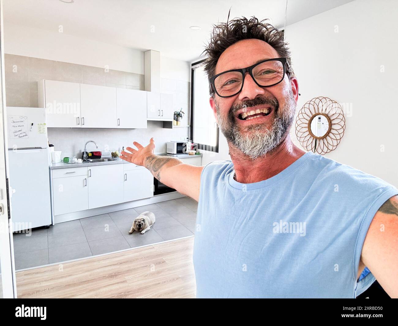 Ein Mann dow-Besitzer macht ein Selfie-Foto im Haus, das Küche und Hintergrundraum zeigt. Glücklicher reifer Mann mit Bart und Brille lächelt in die Kamera und zeigt die Inneneinrichtung. Fröhliche Leute Stockfoto