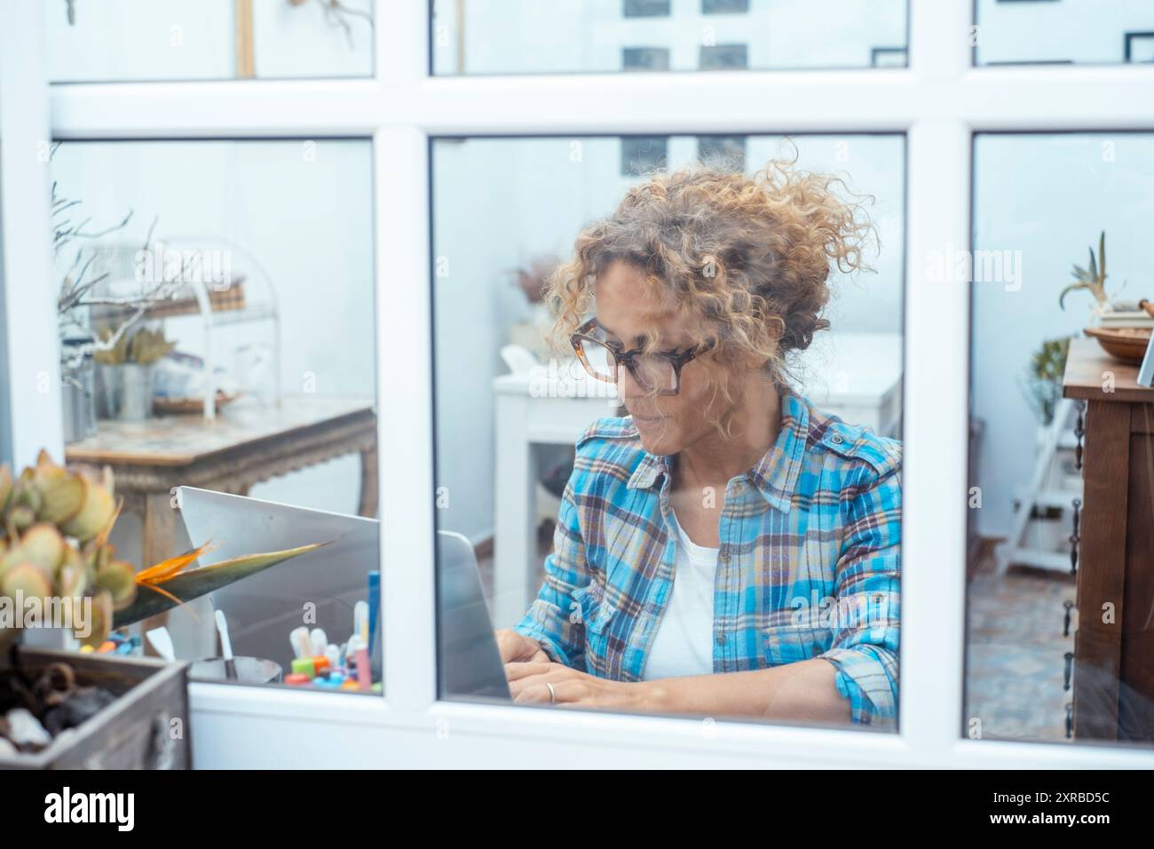 Frau in kariertem Hemd, die am Laptop arbeitet, während sie am Schreibtisch in der Nähe des Fensters in einem Laborbüro zu Hause sitzt. Sicht durch das Fenster. Kleine neue Unternehmen, weibliche Menschen, die ihren Computer online nutzen Stockfoto