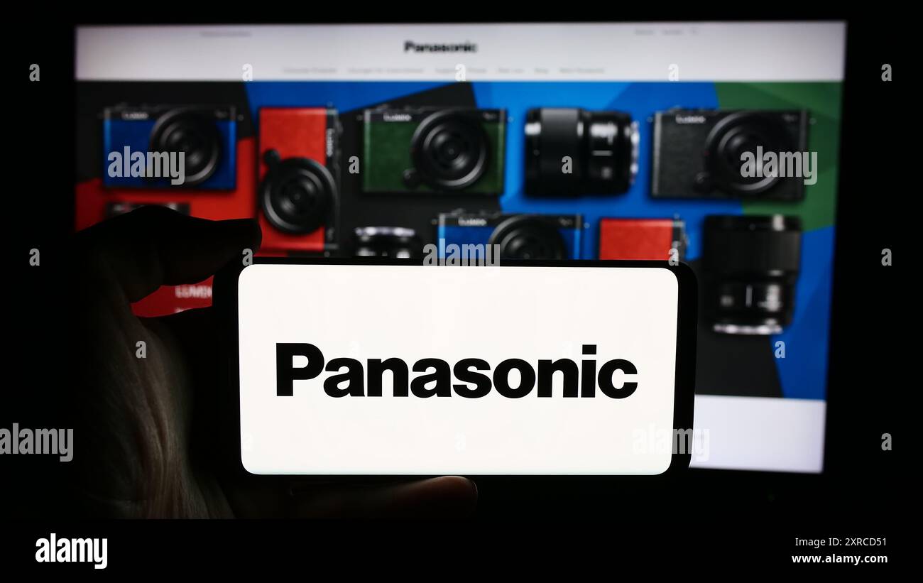 Person, die ein Mobiltelefon mit dem Logo der japanischen Firma Panasonic Holdings Corporation vor der Webseite hält. Schwerpunkt auf dem Display des Telefons. Stockfoto
