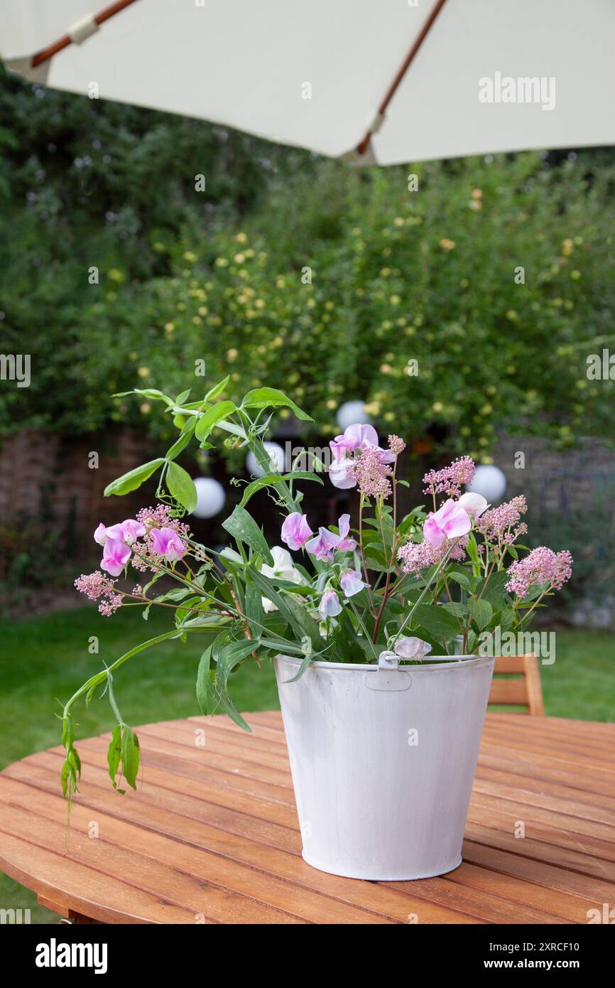 Ein weißer Cachepot mit frischen Blumen wie rosa Sackblumen, weißen und rosa Wicken und grünen Blättern steht als Blumenarrangement auf einem Holztisch im Garten, ein Apfelbaum mit hellgrünen Äpfeln steht verschwommen im Hintergrund Stockfoto