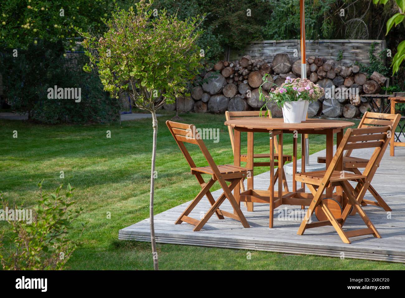 Gartenmöbel auf einer Holzterrasse im letzten Sonnenlicht des Tages, Sitzen in der Abendsonne im grünen Garten im Spätsommer, ein kleiner Fliederbaum im Vordergrund Stockfoto