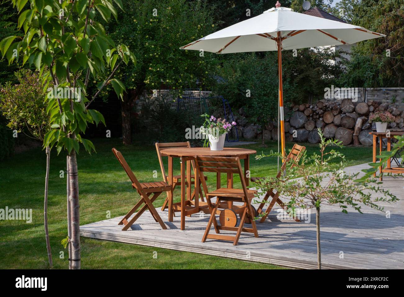 Gartenmöbel mit Sonnenschirm auf einer Holzterrasse im letzten Sonnenlicht des Tages, Sitze in der Abendsonne im grünen Garten im Spätsommer, im Vordergrund ragen die grünen Blätter einer Weide und eines Kirschbaumes ins Bild Stockfoto