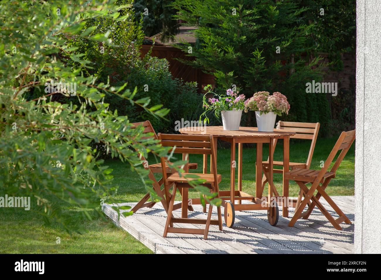 Gartenmöbel auf einer Holzterrasse im letzten Sonnenlicht des Tages, Sitze in der Abendsonne im grünen Garten im Spätsommer ragen die grünen Blätter einer Weide im Vordergrund ins Bild Stockfoto