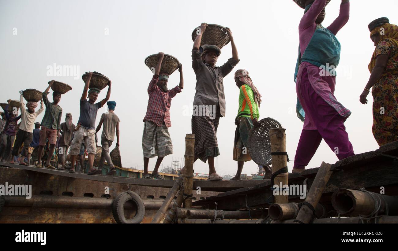 Eine menschliche Kette von Trägern trägt Kohle, Sand und Kies von den Bargen, die an der Aminbazar Landing Station am Buriganga River außerhalb von Dhaka festgemacht sind. Bangladesch absolviert derzeit den Abschluss der LDC-Kategorie (am wenigsten entwickelte Länder), was zum großen Teil der extrem harten Arbeit billiger Arbeitskräfte zu verdanken ist. Ein Portier macht zwischen 80 und 140 USD pro Monat, laut den Websites paylab.com und averagesalarysurvey.com Stockfoto
