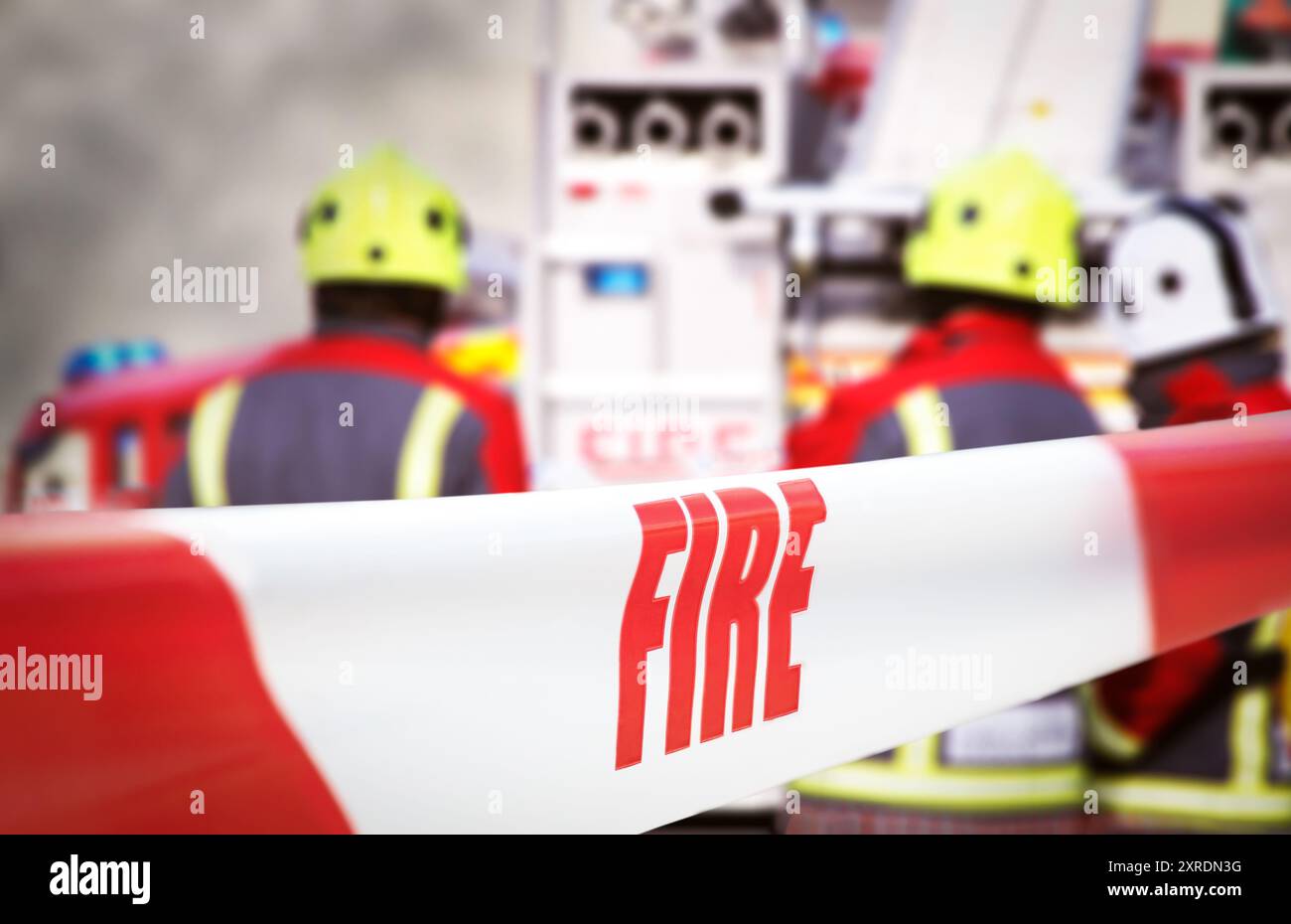 Mit rotem und weißem Band abgeriegelt, mit der Aufschrift „Feuer“. Im Hintergrund sind drei Feuerwehrleute und zwei Feuerwehrmaschinen unscharf Stockfoto