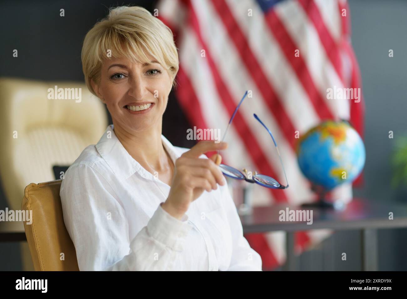 Eine Frau mit einer kurzen blonden Frisur sitzt selbstbewusst an ihrem Schreibtisch, hält ihre Brille und lächelt mit der amerikanischen Flagge hinter sich. Stockfoto
