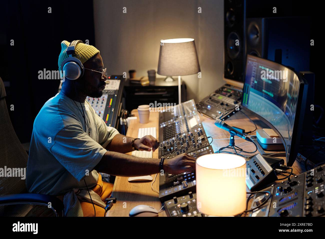 Mann, der an der Mischkonsole im Studio sitzt und den Audiopegel mit Konzentration anpasst. Umgeben von professioneller Audioausstattung und Computerbildschirmen, Kopfhörern und zielgerichtetem Ausdruck Stockfoto