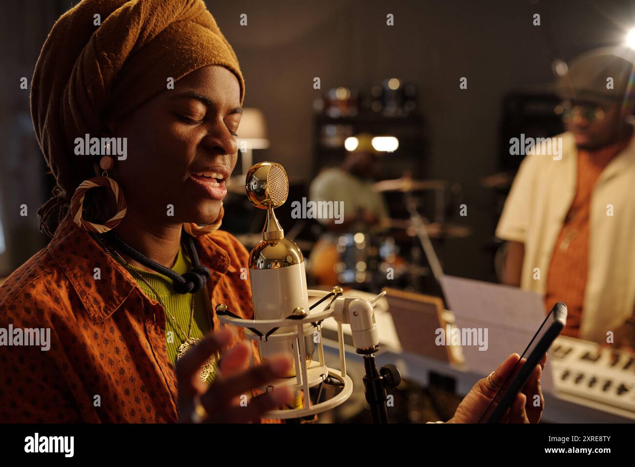 Afroamerikanerin singt in der Nähe des Mikrofons bei der Aufnahme im Musikstudio in schwach beleuchteten Räumen, während Musiker im Hintergrund Instrument spielt Stockfoto