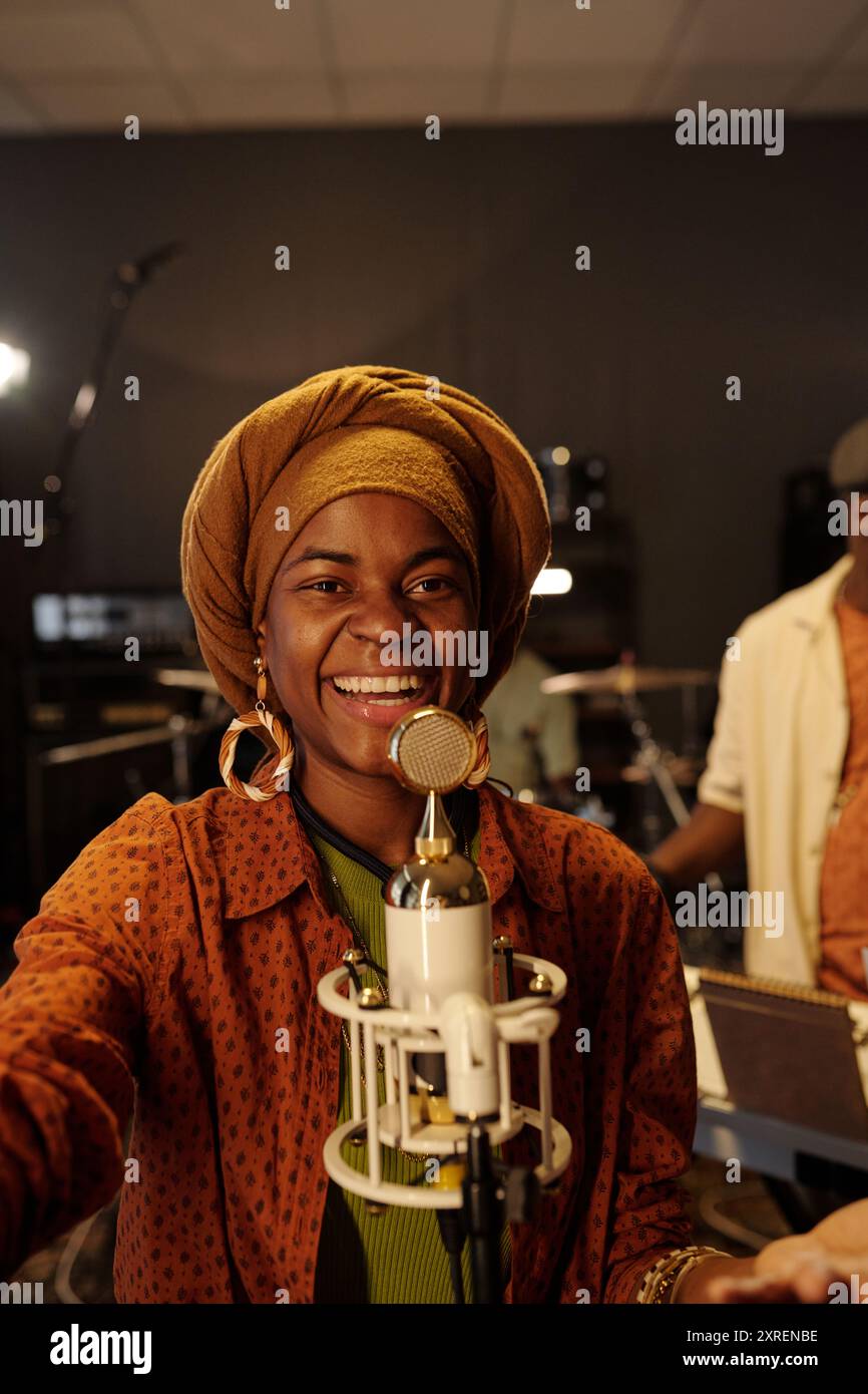 Lächelnde Frau mit Kopftuch und Mikrofon im Musikstudio mit sichtbaren Geräten und Hintergrundelementen Stockfoto