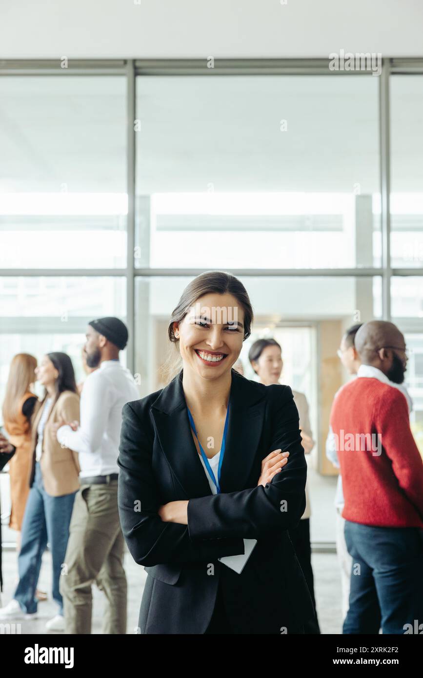 Erfolgreiche Geschäftsfrau lächelt selbstbewusst bei einer Unternehmenskonferenz mit Kollegen, die im Hintergrund vernetzt werden. Stockfoto