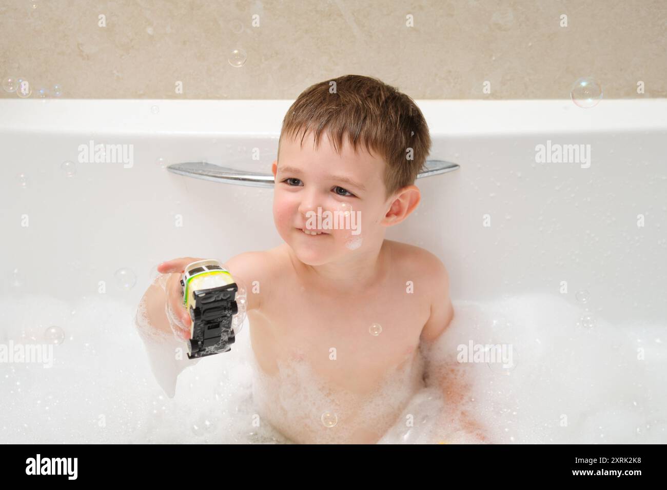 Ein kleiner Junge spielt mit einem Spielzeugauto in der Badewanne. Er lächelt und hält das Spielzeugauto in der Hand. Die Szene ist verspielt und unbeschwert, mit t Stockfoto