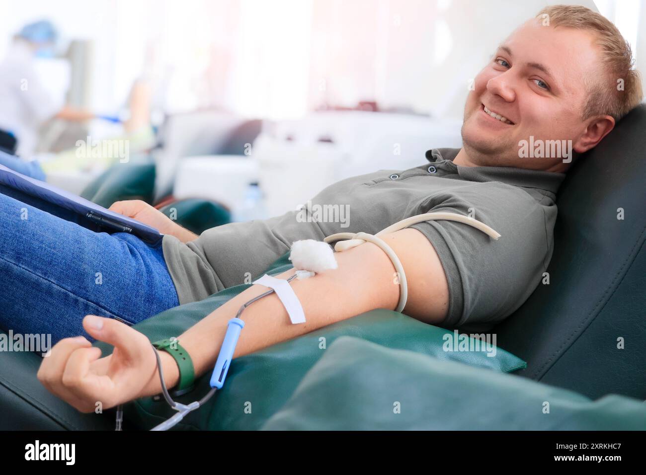 Ein junger Mann sitzt bequem in einer medizinischen Einrichtung und spendet Blut während einer öffentlichen Veranstaltung. Er lächelt, während ein Arzt in der Nähe Geräte vorbereitet. Stockfoto