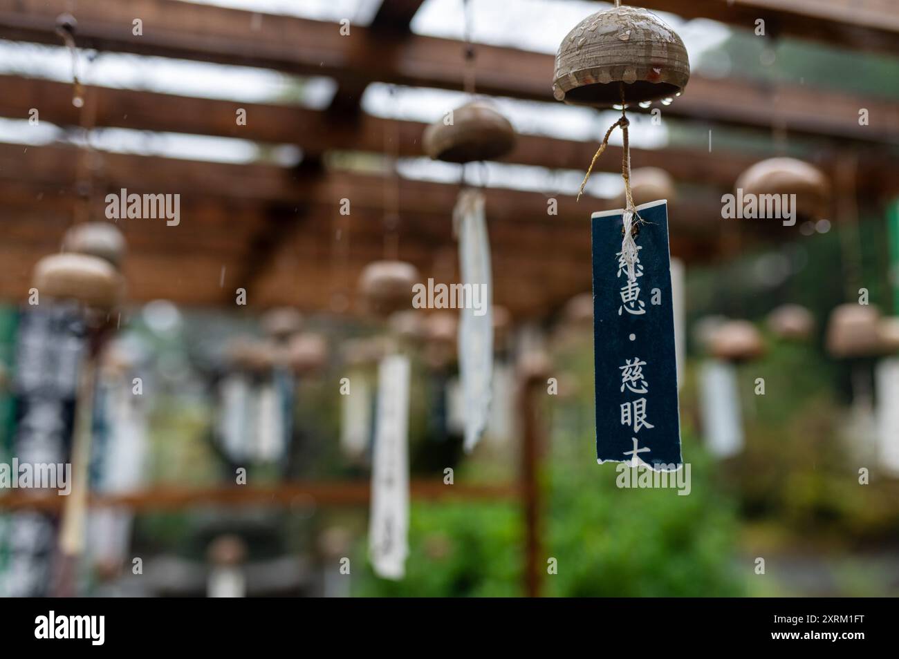 Nahaufnahme traditioneller japanischer Windspiele, die unter einer hölzernen Pergola in einer ruhigen Gartenumgebung hängen, mit Regentropfen für eine friedliche Atmosphäre. Stockfoto