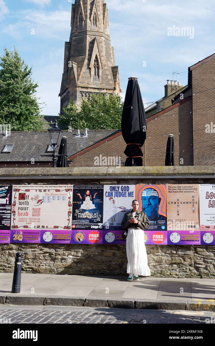Frau steht neben einer Wand, die mit Werbeplakaten für das Fringe Festival in Edinburgh, Schottland, bedeckt ist. August 2024 Stockfoto