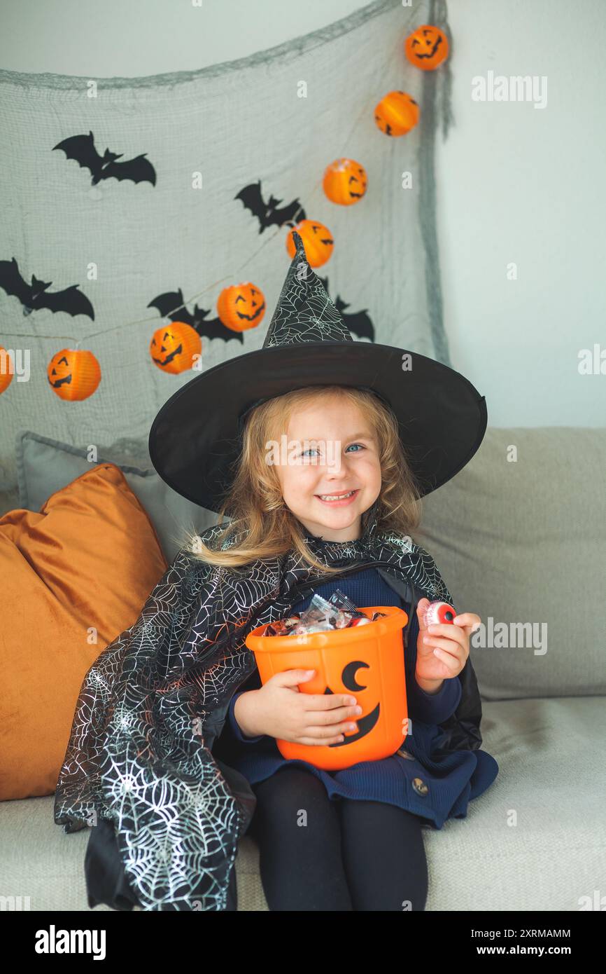 Das kleine süße Mädchen 3 Jahre alt in einem Hexenkostüm mit einem Kürbiseimer voller Süßigkeiten feiert Halloween zu Hause mit Deko. Feiertage. Stockfoto