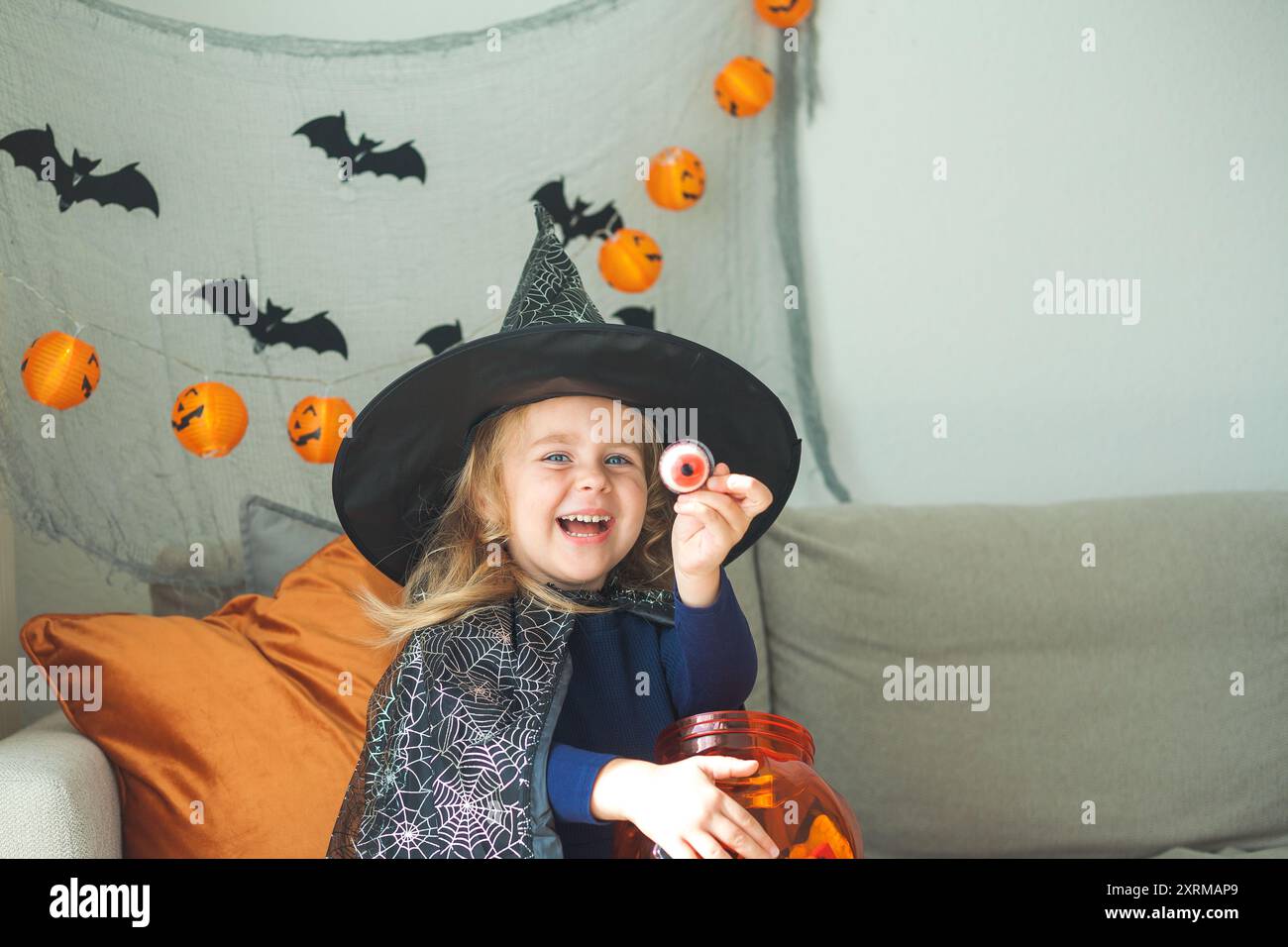 Das kleine süße Mädchen 3 Jahre alt in einem Hexenkostüm mit einem Kürbiseimer voller Süßigkeiten feiert Halloween zu Hause mit Deko. Feiertage. Stockfoto