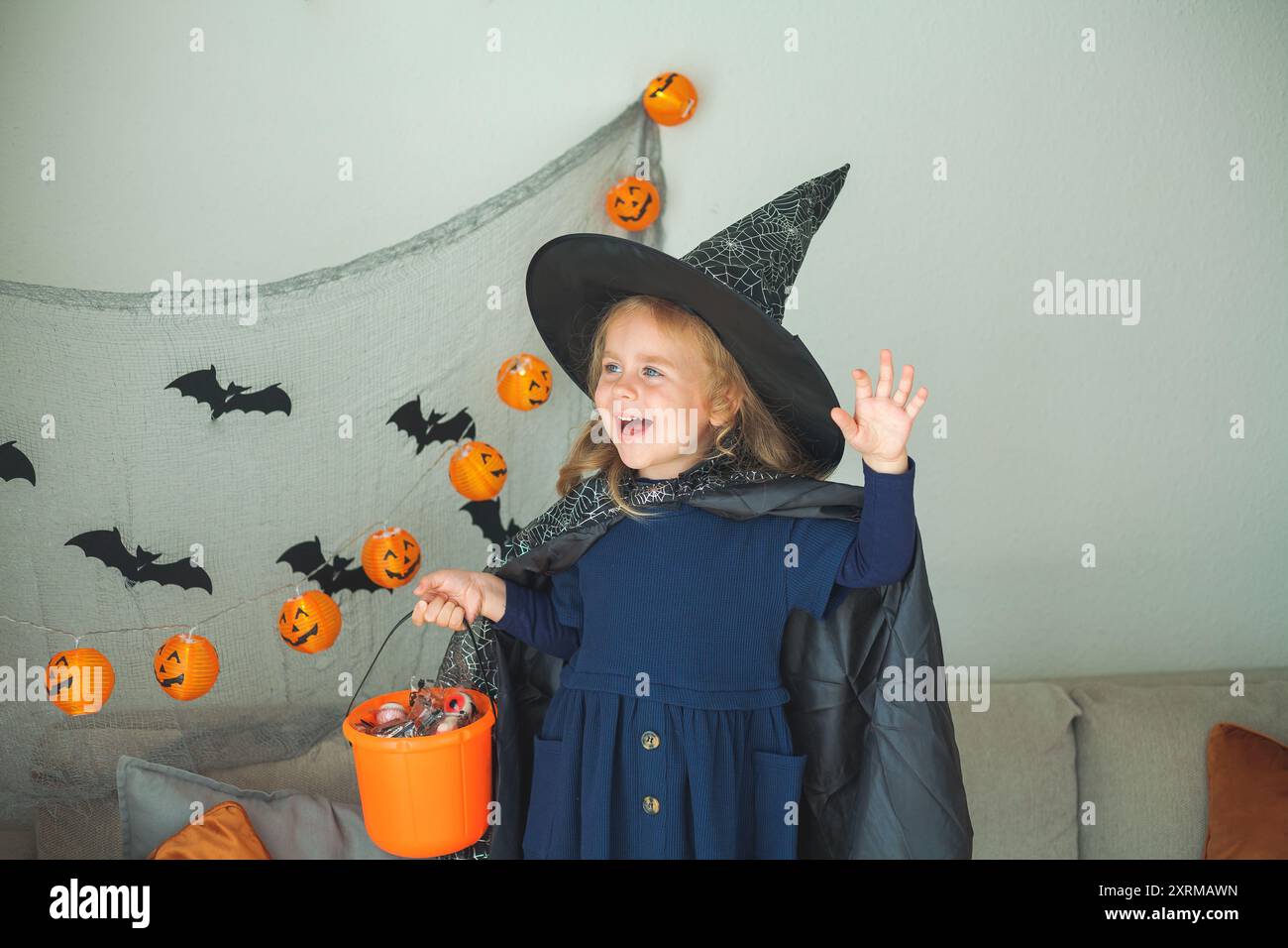 Das kleine süße Mädchen 3 Jahre alt in einem Hexenkostüm mit einem Kürbiseimer voller Süßigkeiten feiert Halloween zu Hause mit Deko. Feiertage. Stockfoto