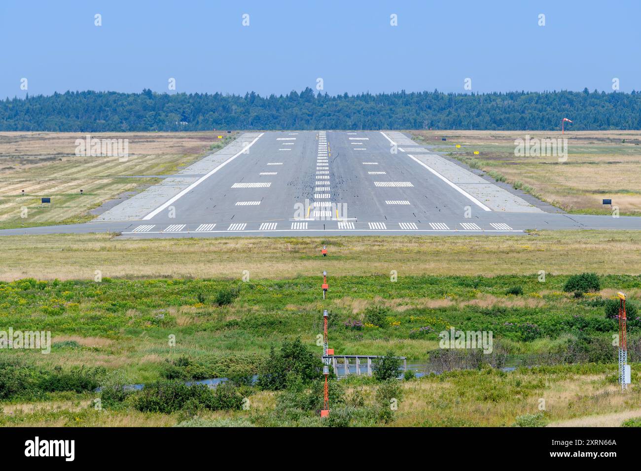 An einem sonnigen Tag erstreckt sich eine Landebahn an einem Flughafen in die Ferne. Feld vor der Start- und Landebahn, Bäume dahinter. Keine Flugzeuge vorhanden, Start- und Landebahn ist leer. Stockfoto