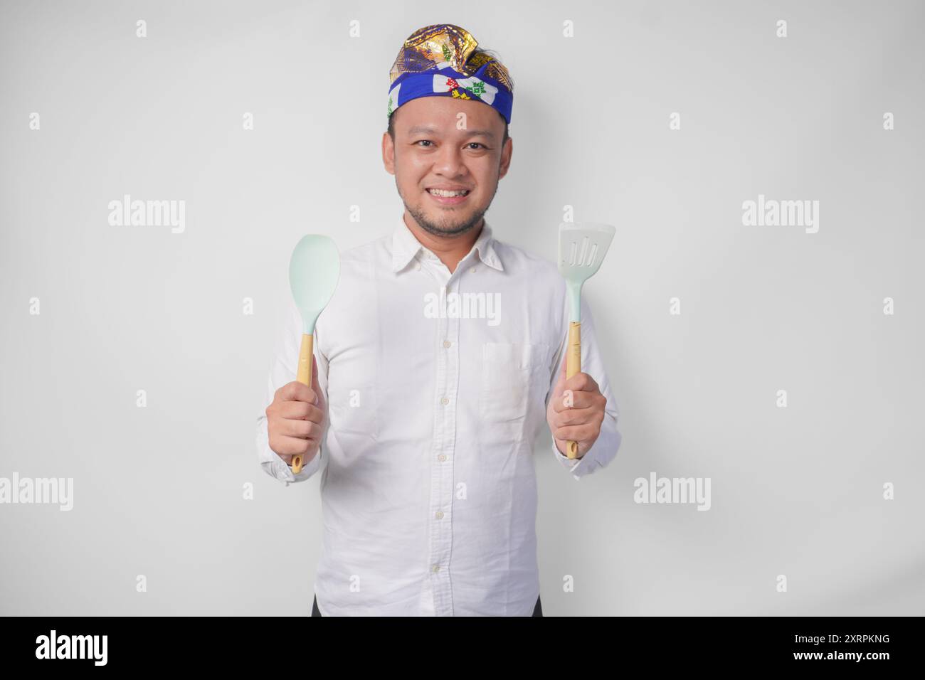 Lächelnder junger balinesischer Mann in weißem Hemd und traditionellem Kopfschmuck mit Spachtel und Küchenutensilien Stockfoto