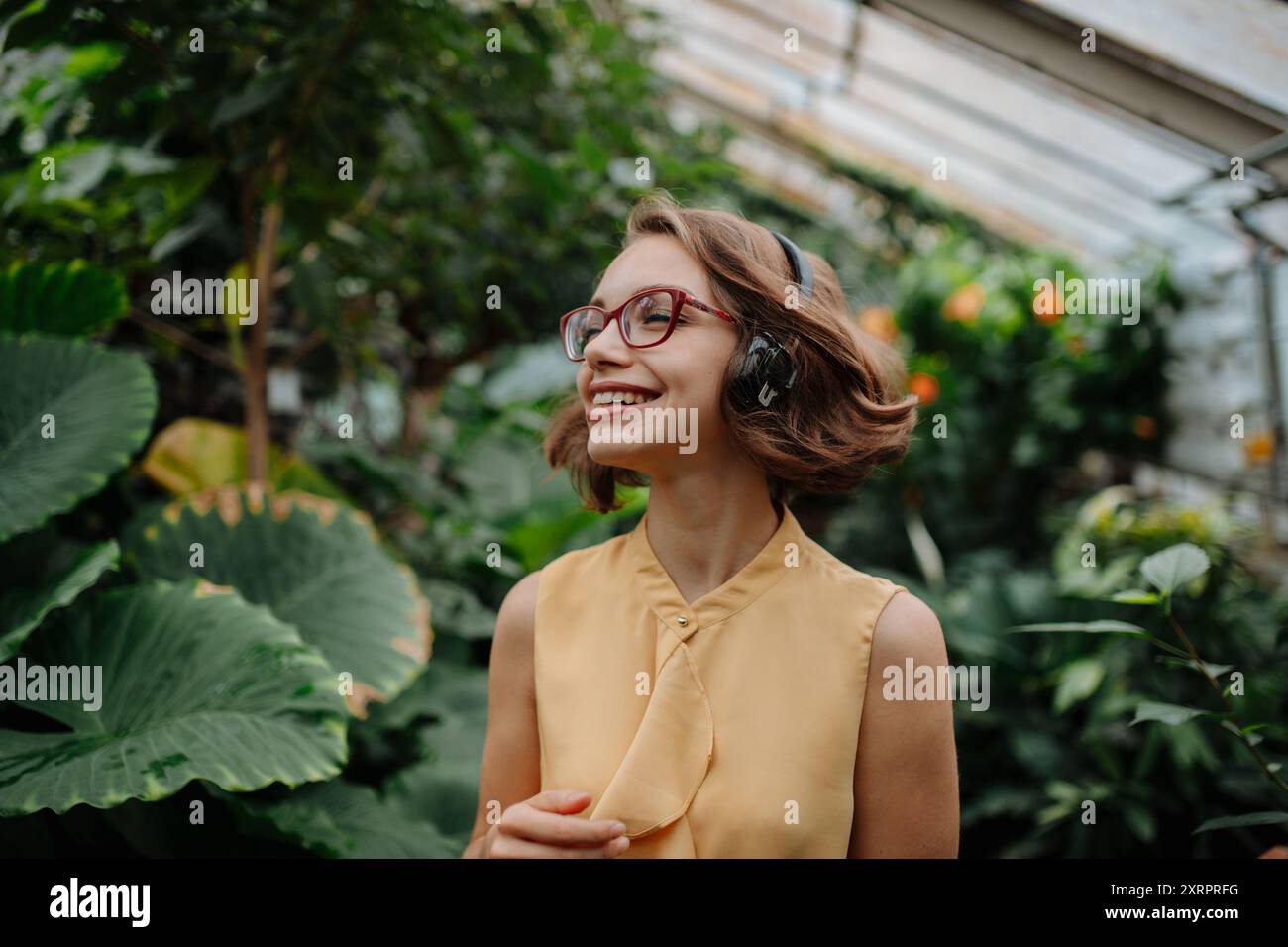 Schöne Frau, die im botanischen Garten steht, mitten in üppig grünem Laub mit Kopfhörern auf dem Kopf, Musik hört. Stockfoto