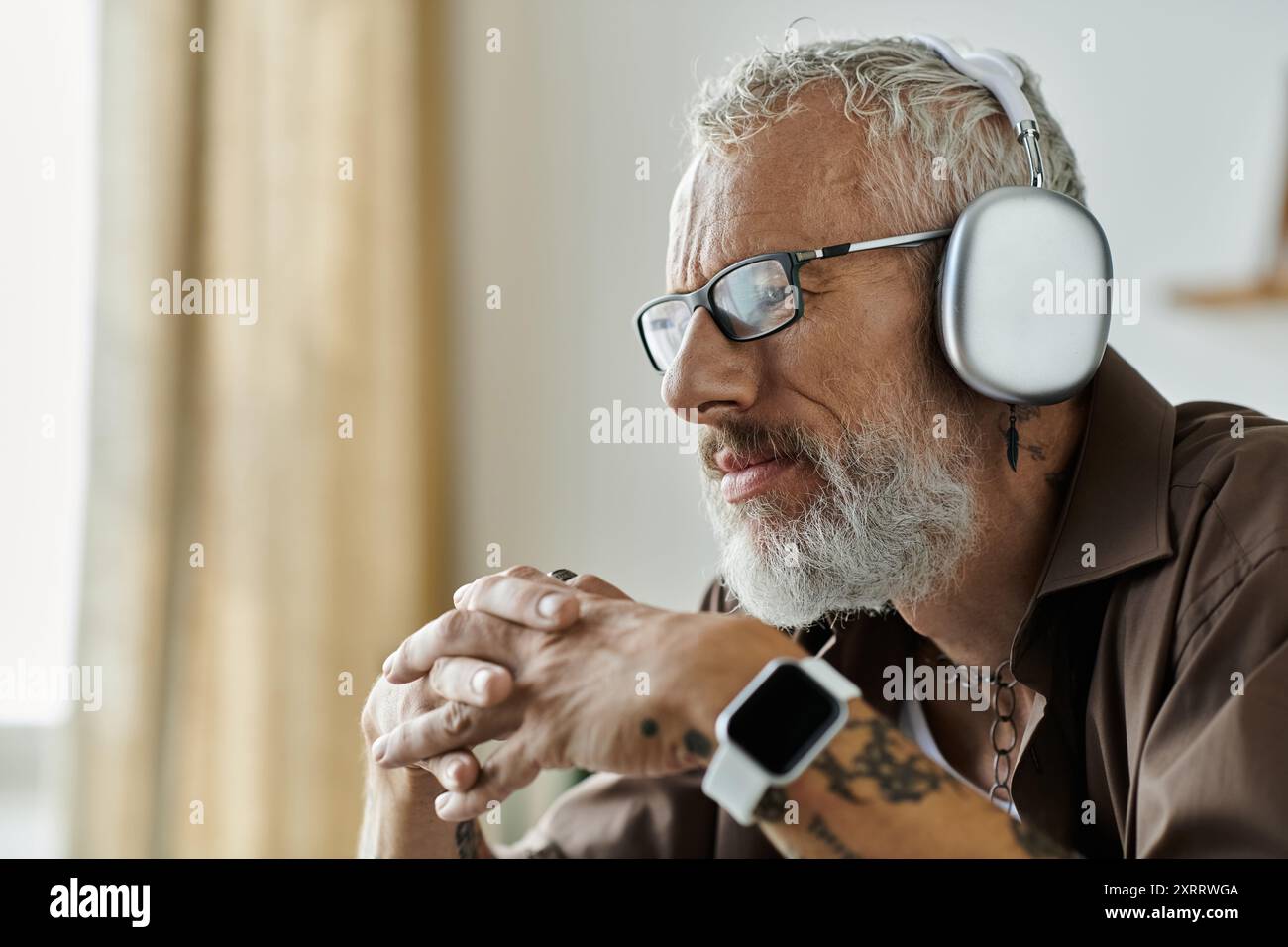Ein reifer schwuler Mann mit Tattoos und grauem Haar arbeitet von zu Hause aus. Er trägt Brille und Kopfhörer. Stockfoto