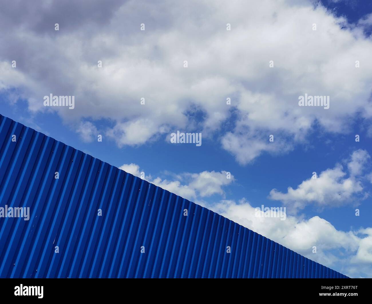 Blaue Metallplatte vor blauem bewölktem Himmel. Anschlussgleise. Nahtlose Oberfläche aus verzinktem Stahl. Industriebauwand aus Wellblech, flach Stockfoto