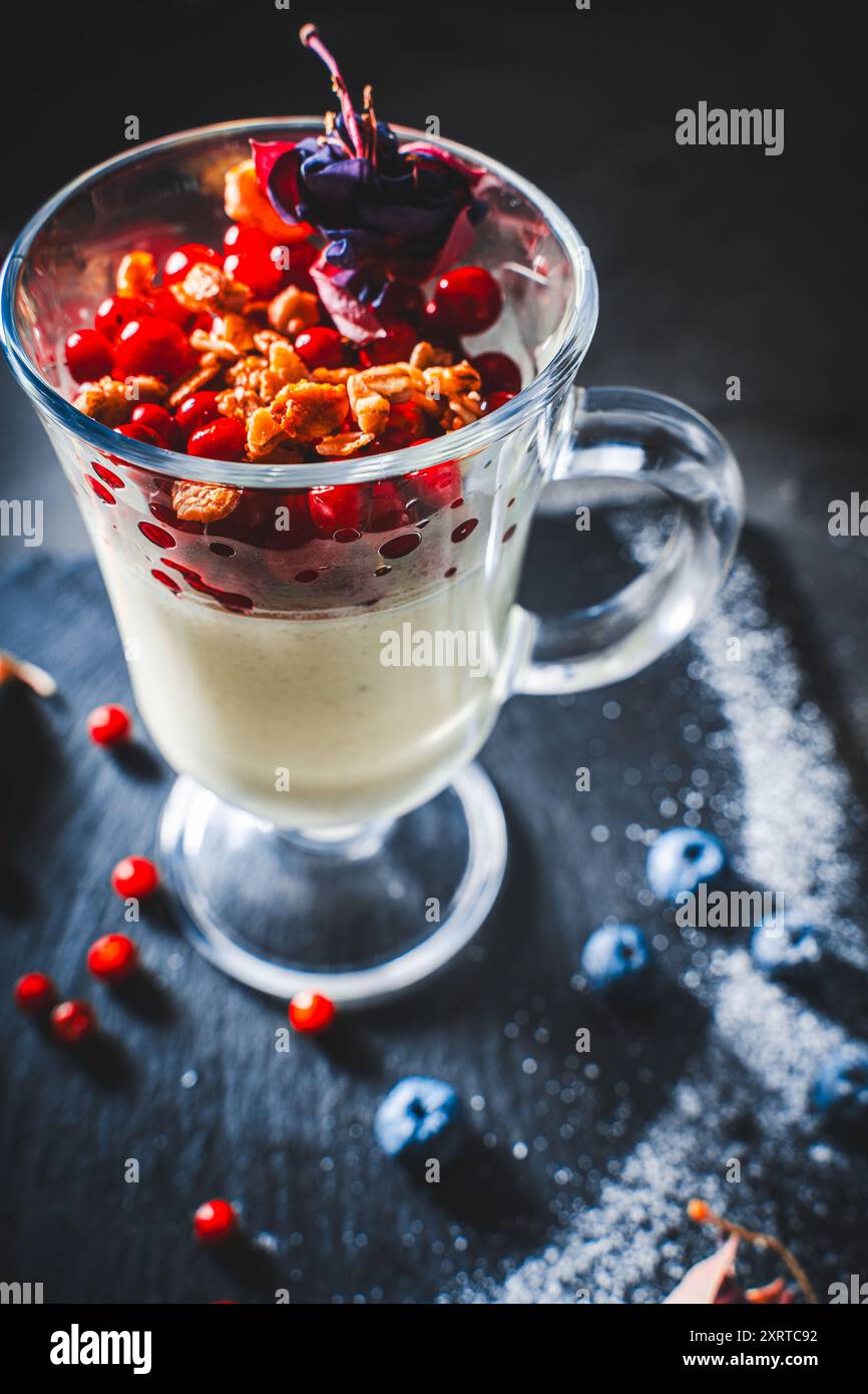 Panna Cotta mit Preiselbeeren und Granola, dekoriert mit getrockneten Blumen auf schwarzem Hintergrund. Ein leichtes und gesundes, diätetisches Dessert. Vertikales Foto. Stockfoto